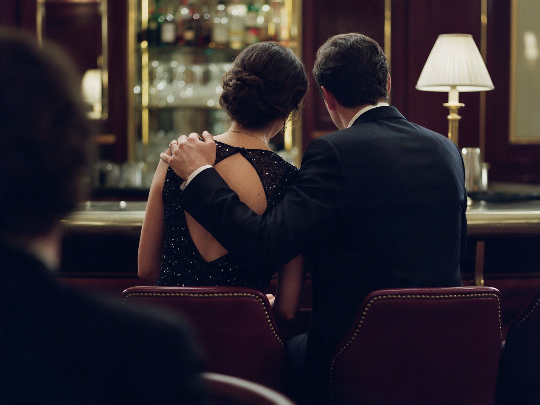 Un couple de dos au comptoir d’un bar d’hôtel, la main de l’un posée sur l’épaule de l’autre
